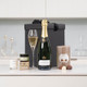 Pampering Treat Box With Champagne. A bottle of champagne is placed next to a filled champagne flute on a white countertop. Beside them are gourmet chocolates and a small jar. In the background, there is a black gift box with a ribbon. The setting features minimalist kitchen decor. Pampering Treat Box With Champagne. A bottle of champagne is placed next to a filled champagne flute on a white countertop. Beside them are gourmet chocolates and a small jar. In the background, there is a black gift box with a ribbon. The setting features minimalist kitchen decor.